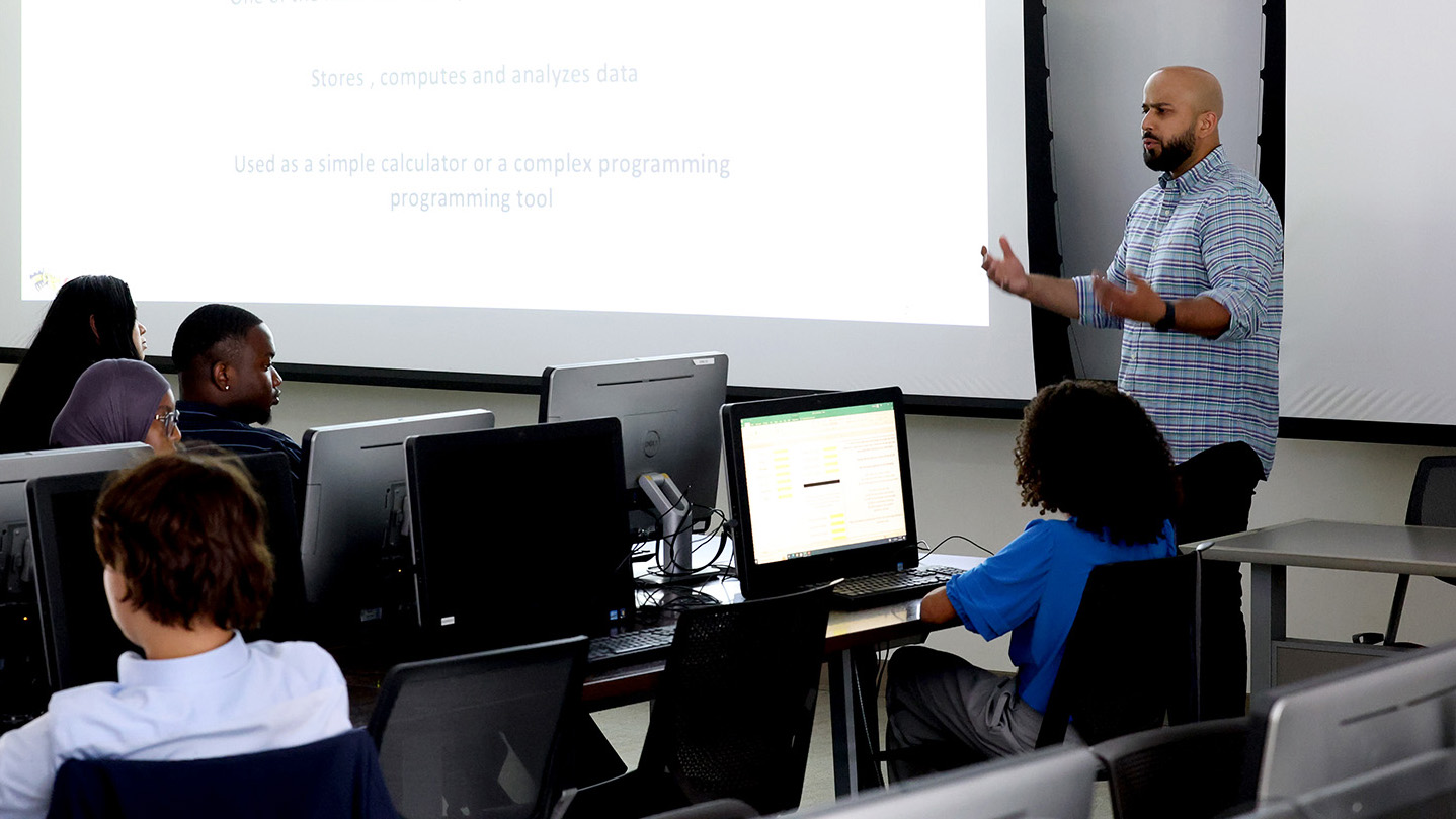 A professor teaches at the front of the classroom in front of a screen