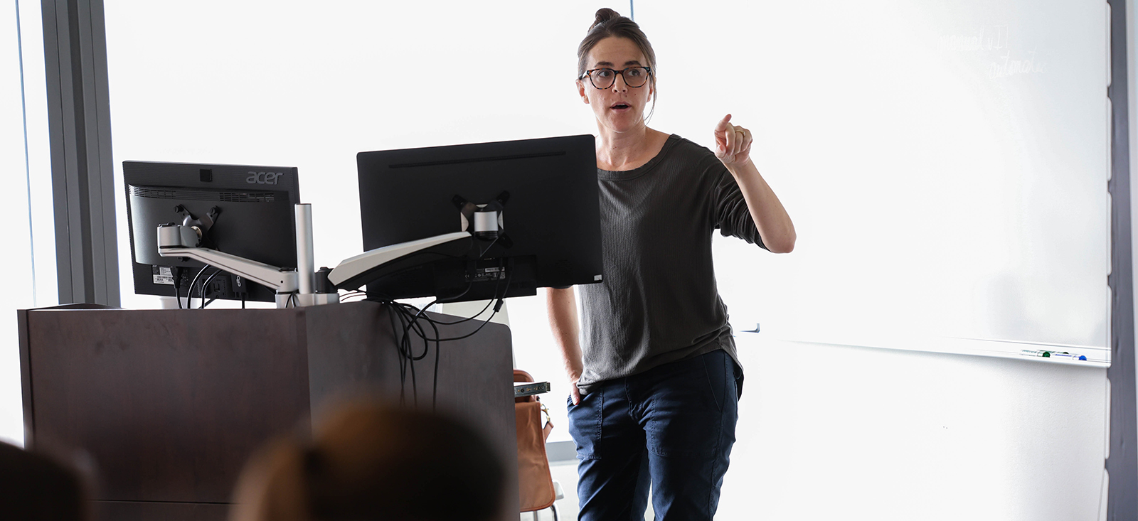 A woman with brown hair and glass pointing with her left hand and standing behind two computer monitors