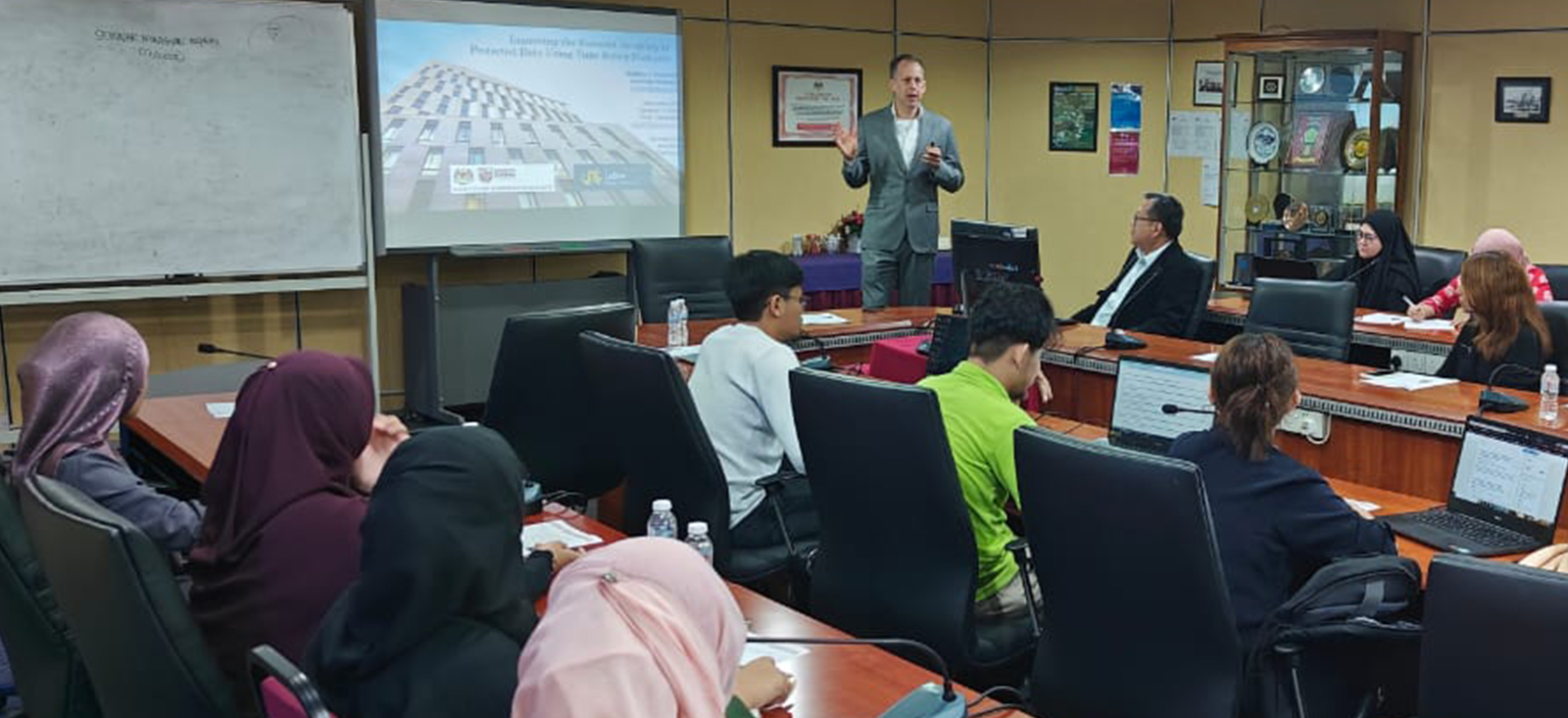 A man in a suit stands in front of students in a classroom with a presentation slide visible in the background