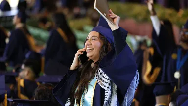 A female student in a blue graduation cap and gown waving to her family while talking on the phone