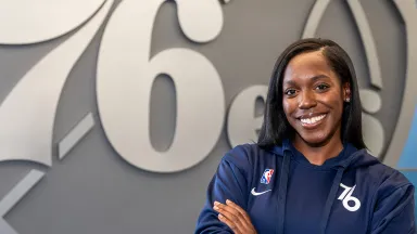 A young Black woman in a blue sweatshirt with the Philadelphia 76ers logo, standing in front of a large silver rendition of the 76ers logo and with her arms crossed in front of her chest