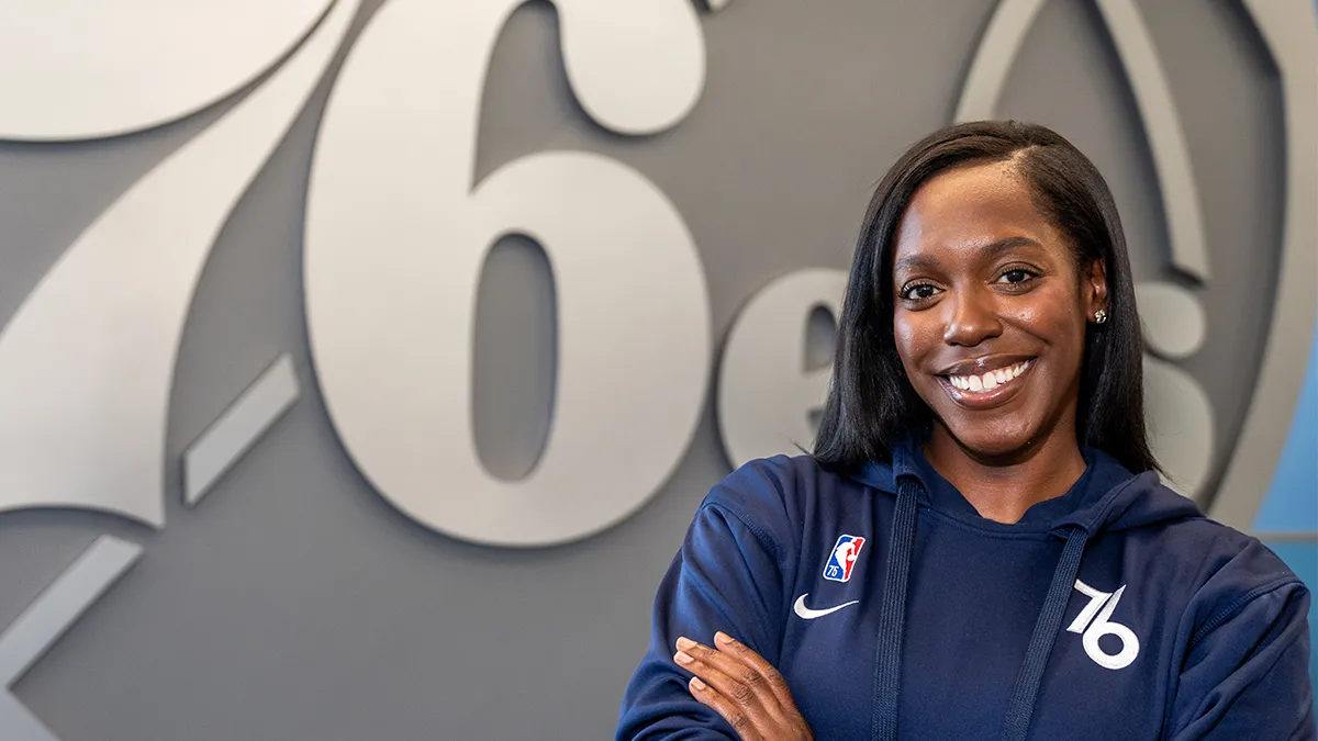 A young Black woman in a blue sweatshirt with the Philadelphia 76ers logo, standing in front of a large silver rendition of the 76ers logo and with her arms crossed in front of her chest